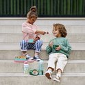 Enfants assis sur des marches. Une fille en pantalon rayé nourrit un garçon avec un bol vert et des raisins.