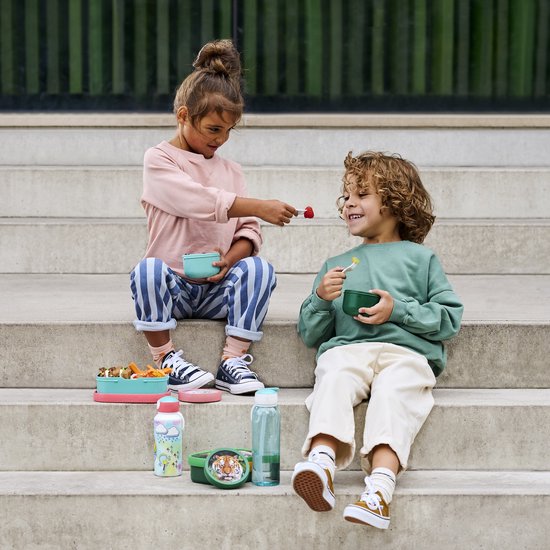 Enfants assis sur des marches. Une fille en pantalon rayé nourrit un garçon avec un bol vert et des raisins.