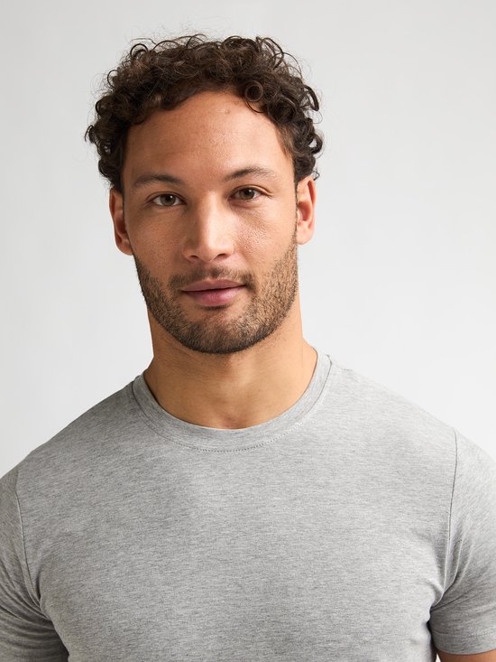 Close-up van een man met krullend haar die een effen lichtgrijs T-shirt met ronde hals draagt.