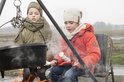 Twee jongens die marshmallows roosteren boven een vuur. Een jongen draagt een oranje jas, de ander een groene jas.