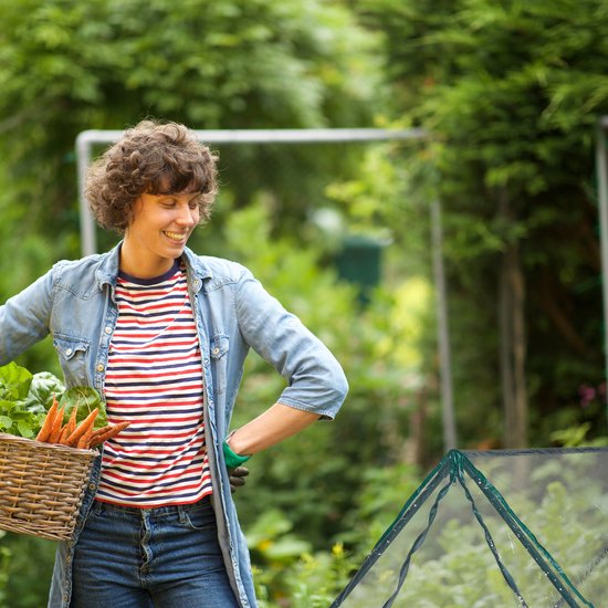 Vrouw met krullend haar in een tuin, draagt een spijkerjasje over een gestreepte top, met een mand met wortels.