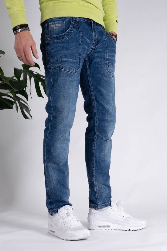 Blauwe spijkerbroek met contrasterende stiksels en een gebruikte wassing. Gedragen door een man met witte sneakers en een limegroene trui.