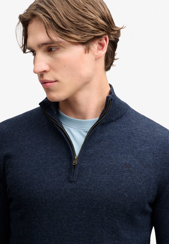 Close-up van een man met donkerblauwe melange trui met ritssluiting en lichtblauw T-shirt eronder.