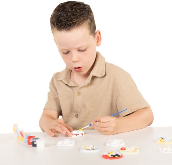 Jongetje met beige shirt schildert met een kwast op een wit gipsen figuur. Diverse andere figuren liggen op tafel.