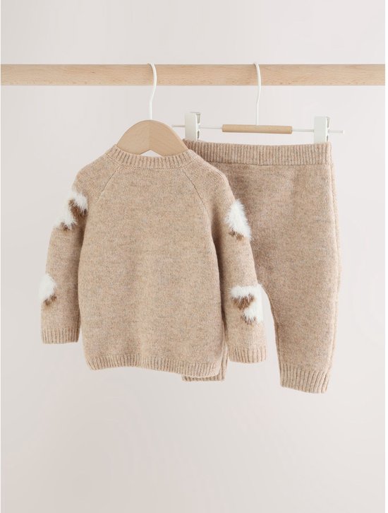 Een beige gebreide babyset met een trui met schaapapplicaties en een bijpassende legging, hangend aan een houten rek.
