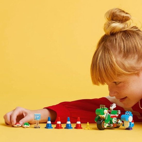 Een kind met een rode trui bouwt aan een groene Lego-motor met een blauwe Yoshi erop. Ernaast staan pionnen en een gele banaan.