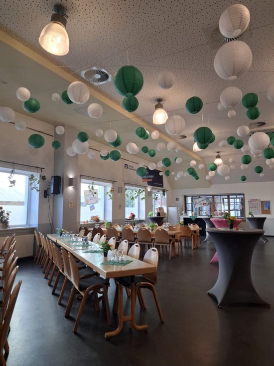 Een zaal met tafels gedekt voor een maaltijd, versierd met witte en groene lampionnen aan het plafond.