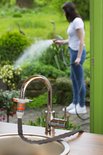 Zilverkleurige kraan met zwarte slang en oranje connector, met een vrouw op de achtergrond die een tuin besproeit.