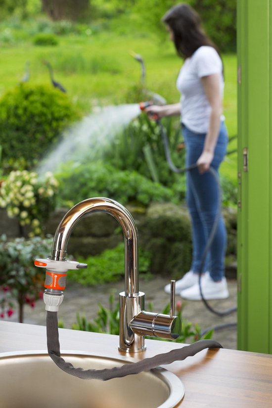 Zilverkleurige kraan met zwarte slang en oranje connector, met een vrouw op de achtergrond die een tuin besproeit.