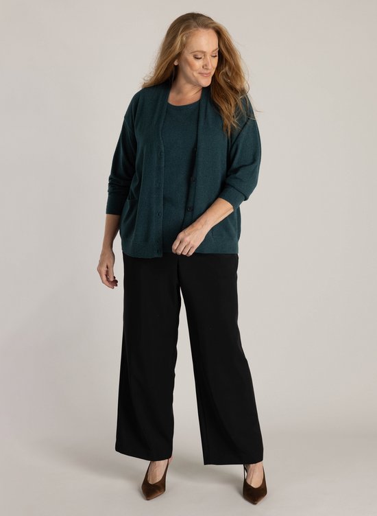 Femme aux longs cheveux roux, porte un gilet tricoté vert foncé boutonnné avec poches plaquées, associé à un caraco et un pantalon noir.