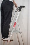 Een persoon staat op een aluminium Baulich trap met antislip treden, donkere broek en witte schoenen.
