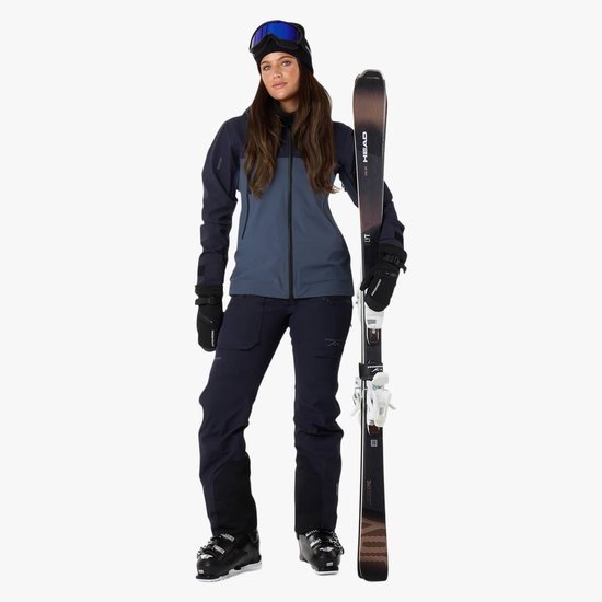 Model in een blauw-grijze ski-jas en donkerblauwe skibroek, met ski's.