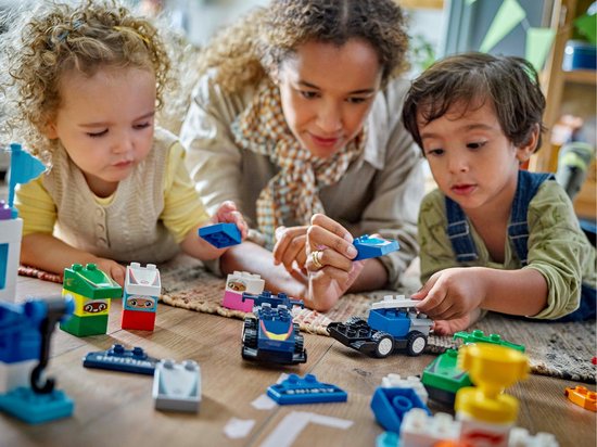 Une mère et deux enfants jouent avec des voitures et des pièces LEGO Duplo bleues sur le sol.