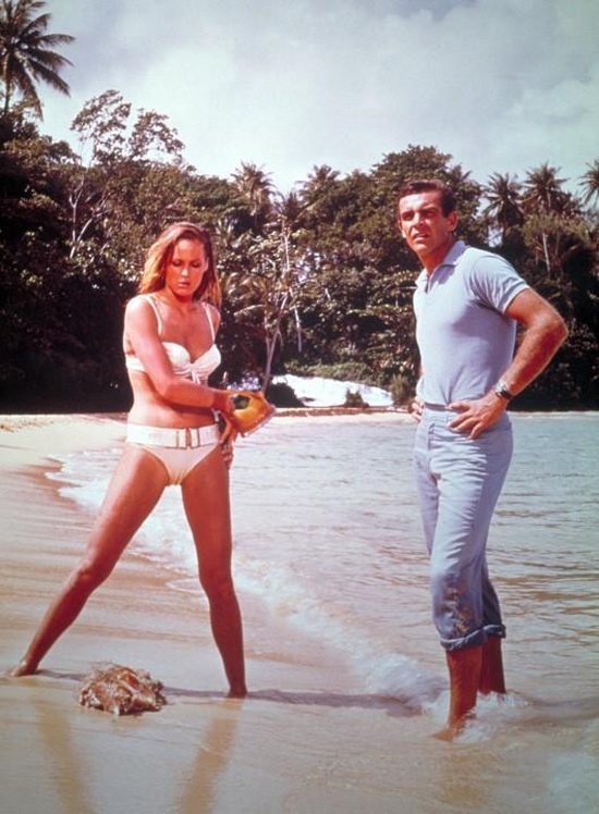 Een vrouw in een witte bikini en een man in een blauw shirt en broek staan op een strand met palmbomen.