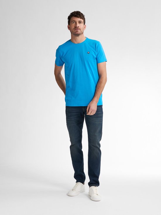Man in blauw t-shirt en spijkerbroek, staand met handen in de zakken.