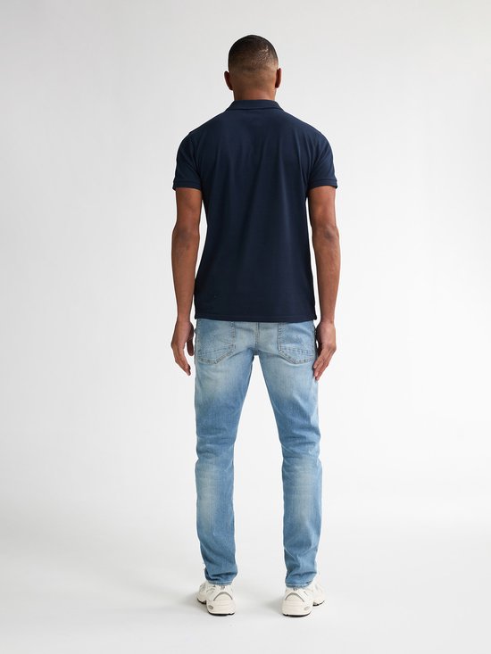 Man, van achteren gezien, draagt een marineblauwe polo en een lichtblauwe slim fit jeans met lichte wassing.