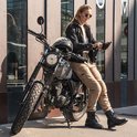 Vrouw met zonnebril, zittend op een motorfiets, draagt een zwarte jas en zandkleurige cargobroek.