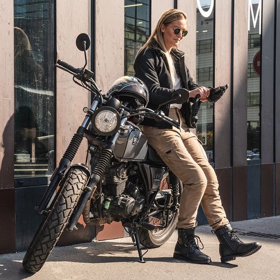 Vrouw met zonnebril, zittend op een motorfiets, draagt een zwarte jas en zandkleurige cargobroek.