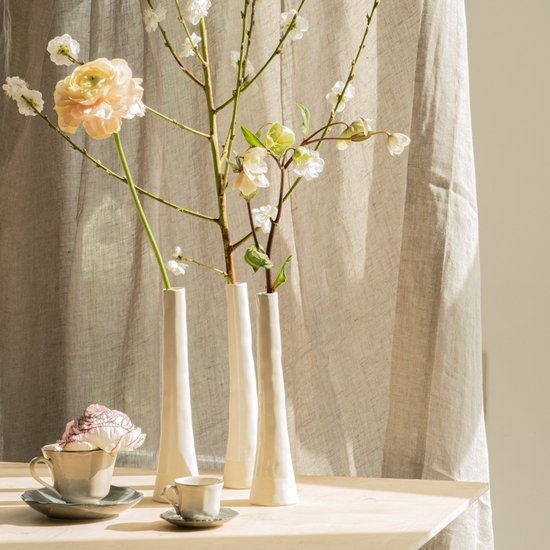 Drie crèmekleurige keramieken vazen op een houten tafel met bloemen, naast twee kopjes.