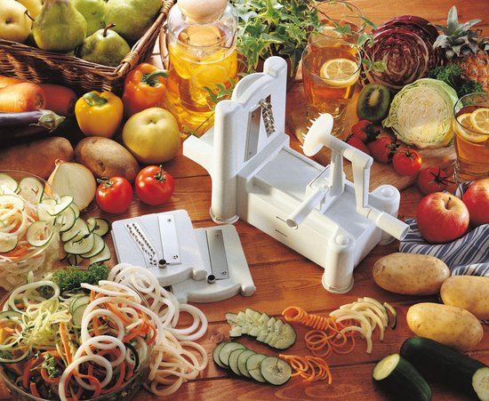 Een witte spiraalsnijder met verschillende snijbladen en verse groenten op een houten tafel.