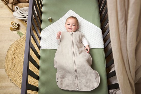 Baby ligt in een wieg in een beige slaapzak, glimlacht naar de camera.