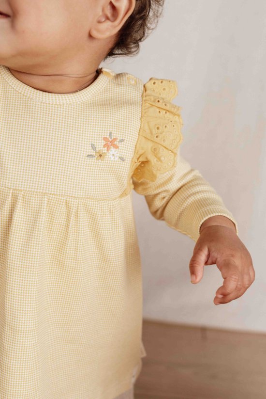 Close-up van een baby in een gele jurk met ruitjes, bloemenborduurwerk en kanten details op de schouders.