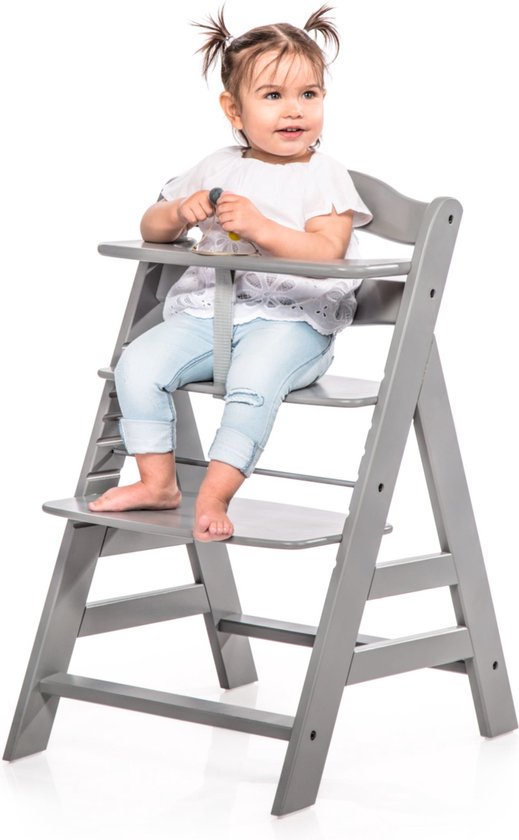 Een jong meisje met staartjes zit in een grijze kinderstoel met een wit kanten shirt en een blauwe spijkerbroek.
