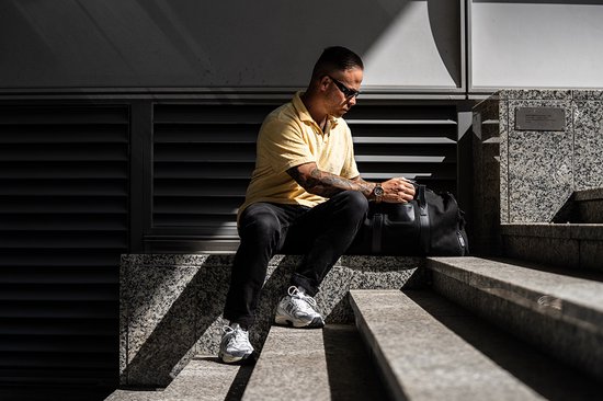 Man met geel poloshirt, zwarte jeans, zittend op een trap met een zwarte sporttas.