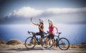 Twee vrouwen met racefietsen maken een selfie bij de zee. De linker vrouw draagt een zwarte fietstop, de rechter een rode.