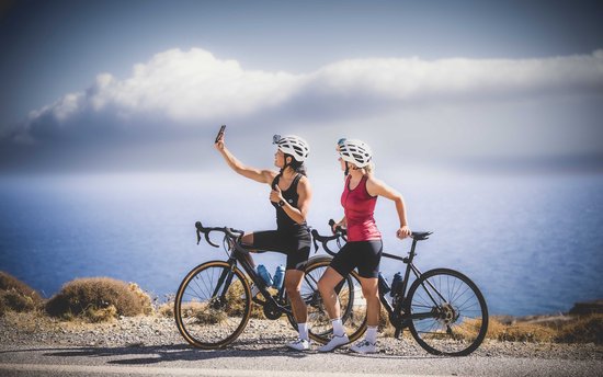 Twee vrouwen met racefietsen maken een selfie bij de zee. De linker vrouw draagt een zwarte fietstop, de rechter een rode.