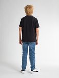 Jongen met blond haar staat van achteren gezien in een zwarte t-shirt en blauwe jeans. Hij draagt witte sneakers.