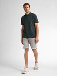 Man van voren gezien, draagt een donkergroene polo en een grijze denim short. Hij draagt witte sneakers met beige accenten.