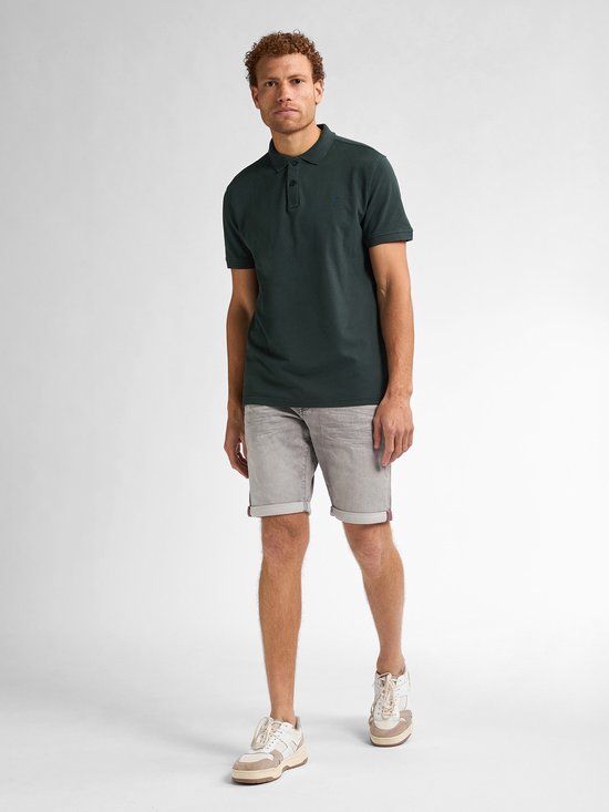 Man van voren gezien, draagt een donkergroene polo en een grijze denim short. Hij draagt witte sneakers met beige accenten.