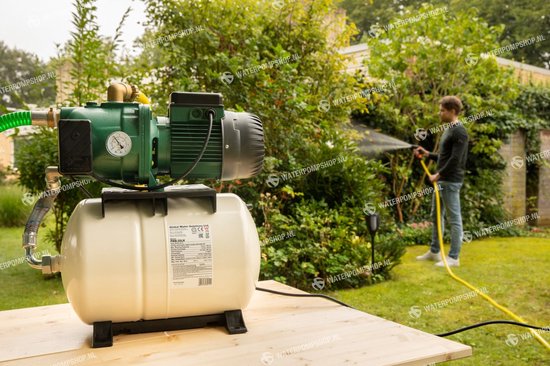 Groene waterpomp met beige druktank op een houten tafel. Een man sproeit met een tuinslang in de tuin.