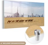 MuchoWow® Glasschilderij 160x80 cm - Schilderij acrylglas - Kamelen die in de woestijn voor Dubai langslopen - Foto op glas - Schilderijen