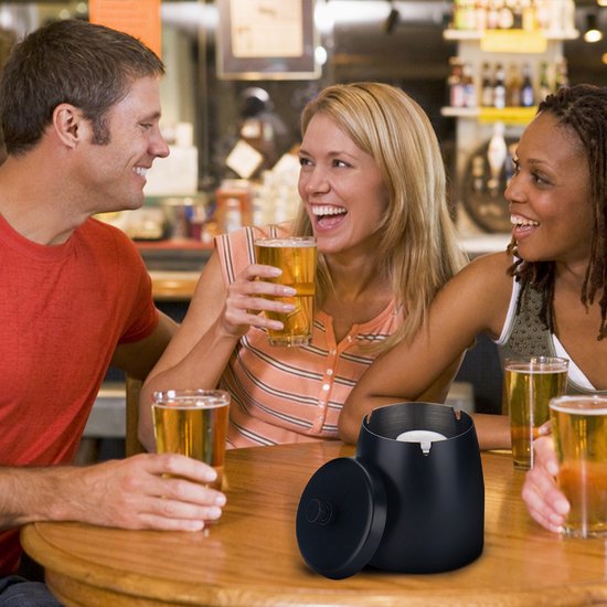 Een zwarte asbak met deksel staat op een tafel in een bar. Drie lachende mensen met bier eromheen.