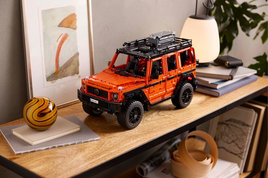 Oranje LEGO Technic Mercedes-Benz G 500 model op een houten tafel naast een lamp en abstracte decoratie.