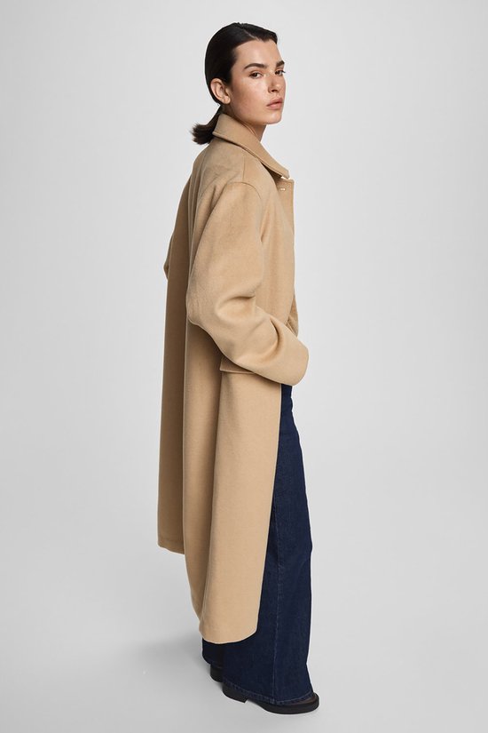 Vrouw in profiel, draagt een lange beige jas met reverskraag en donkere jeans.