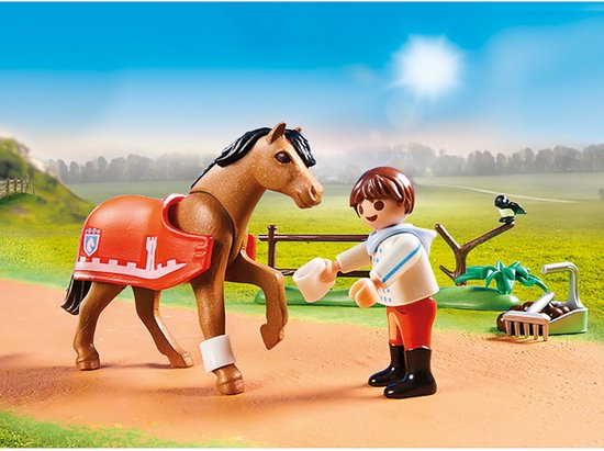 Enfant avec cheval portant une couverture orange avec motif de château. L'enfant tient une mangeoire blanche.