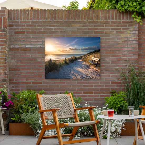 Zonsondergang op een strand met duinen, een zandpad, houten hekken en een bankje. Hangt aan een bakstenen muur in een tuin.