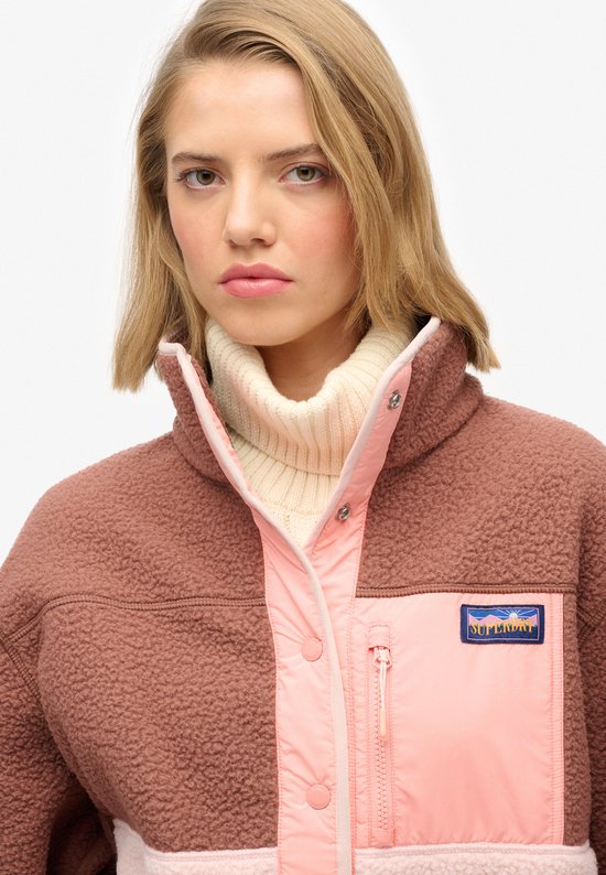 Close-up van de bruine fleece jas met roze en lichtroze panelen en logo.