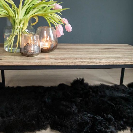Salontafel met licht houten blad en zwart stalen frame. Een boeket roze tulpen en twee glazen windlichten met kaarsen erop. Zwart schapenvachtje eronder.