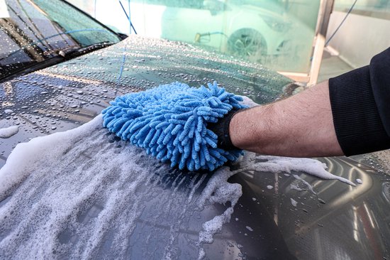 Een hand wordt met een blauwe microvezel washandschoen over een natte, schuimende motorkap bewogen.