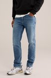 Man met de handen in de zakken, dragend een zwarte sweater en een blauwe jeans met lichte wassing.