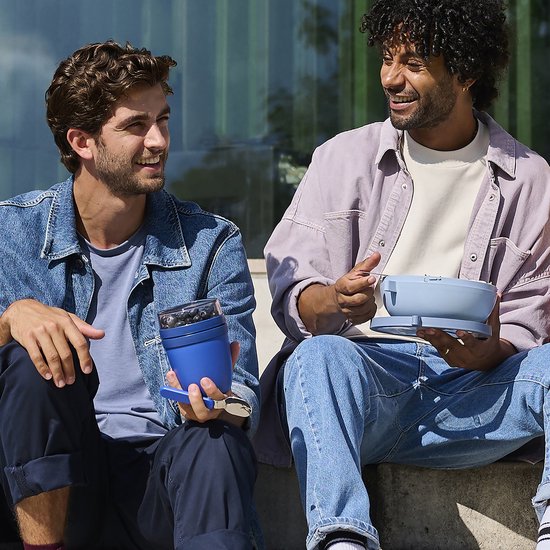 Deux hommes à l'extérieur, l'un tenant un pot à déjeuner et une cuillère bleus, l'autre une boîte à déjeuner bleu clair.