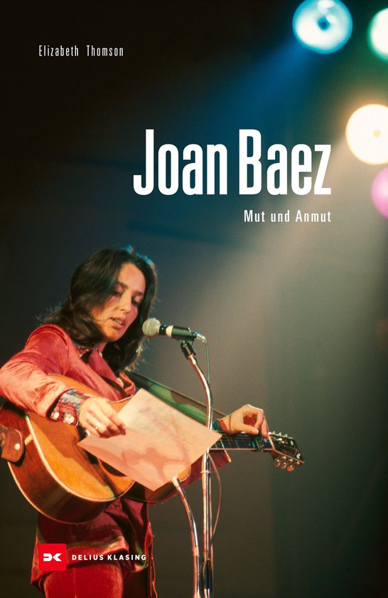 Joan Baez, een vrouw met een gitaar, zingt op een podium onder spotlights.