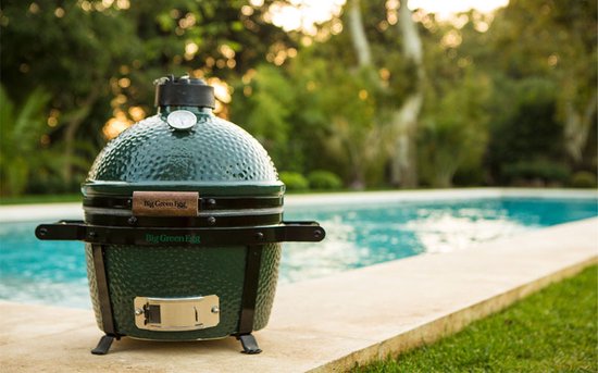 Donkergroene Big Green Egg barbecue met houten handvat en metalen onderstel, bij een zwembad.