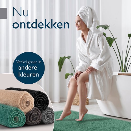 Vrouw in badjas zit naast een bad, naast een stapel gerolde badmatten in verschillende kleuren.