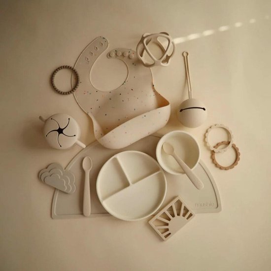 Een crèmekleurige siliconen placemat met daarop een bord met drie vakken, slabbetje, kom, lepels, een babymobiel en een bijtring.