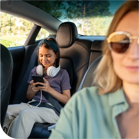 Meisje met koptelefoon kijkt op haar telefoon, zittend op de achterbank van een auto met gordel om.
